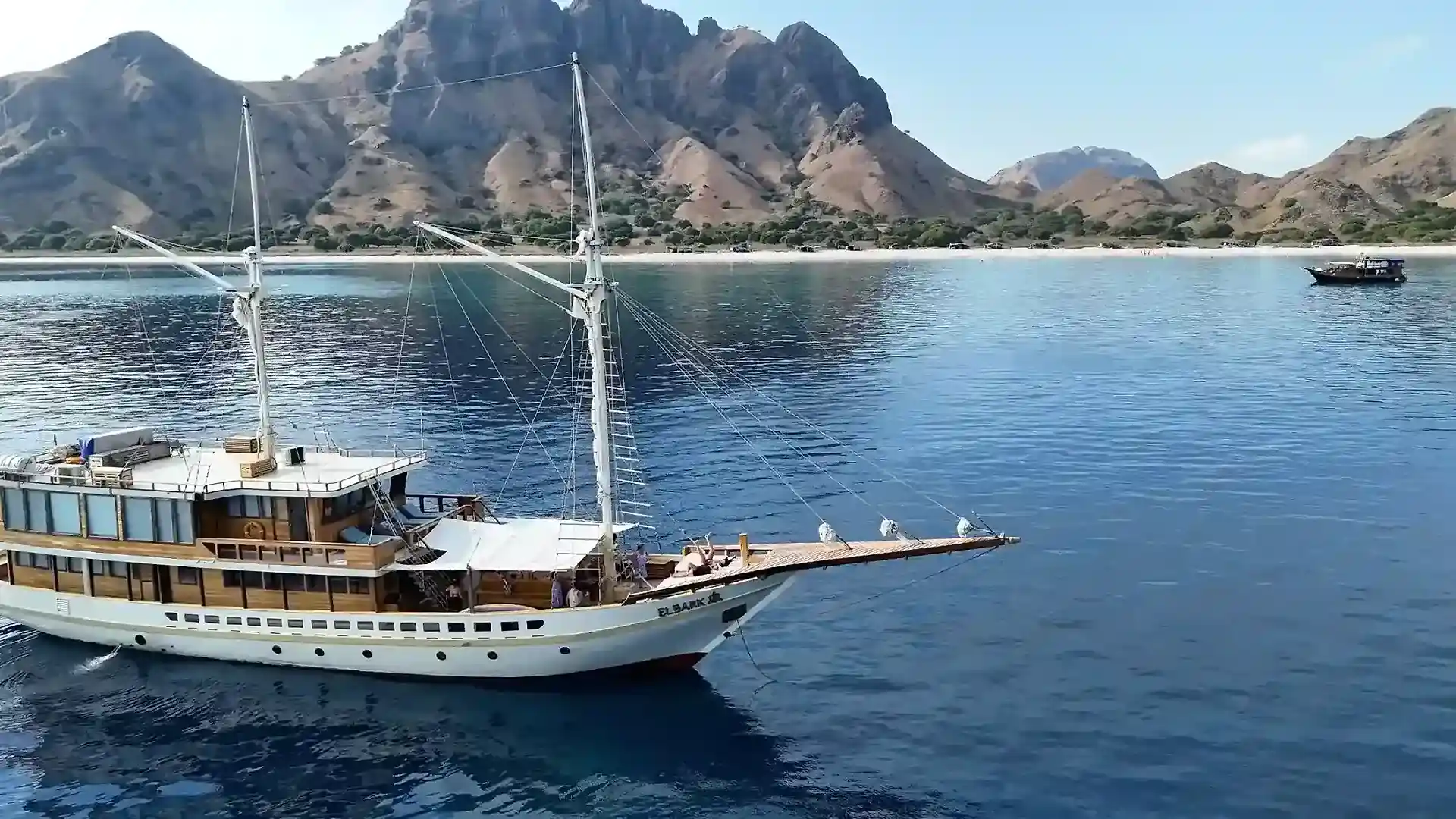 Elbark at anchor in a turquoise bay ringed by green hills